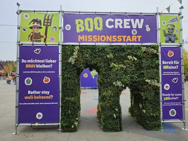 Das BOO CREW Missionsstart-Tor: ein Tunnel aus künstlichem Grün zwischen lila Bannern, die auf Deutsch und Englisch fragen, ob du lieber brav bleiben willst oder bereit für BOO-sinn bist