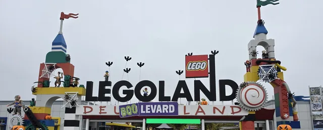 Der Haupteingang des LEGOLAND Deutschland im Halloween-Look: die großen LEGOLAND-Buchstaben mit Fledermäusen auf Stangen darüber, Spinnennetze an den Türmen aus Steinen und ein lila „BOOLEVARD“-Banner über dem Tor
