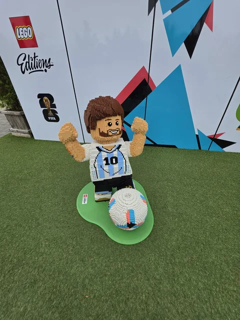 Eine Fußballerfigur aus Steinen im hellblau-weiß gestreiften Trikot mit der Nummer 10, Arme hochgerissen, ein Ball zu seinen Füßen, vor einer LEGO Editions-Kulisse