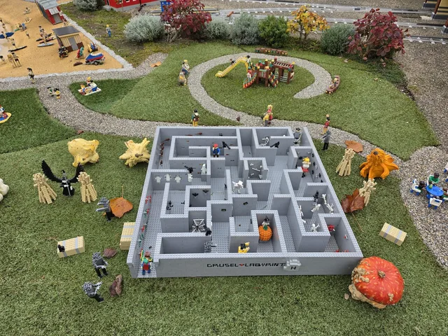 Ein Mini-Labyrinth aus Steinen im Miniland mit der Aufschrift „Grusel-Labyrinth“, voller Skelette, Geister, Spinnennetze und einer Hexe auf einem Kürbis, ringsum echte Kürbisse auf dem Rasen