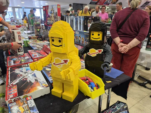 Zwei aus Steinen gebaute Minifiguren in Kniehöhe auf einem Händlertisch — eine gelbe mit Classic-Space-Logo und eine schwarze — umgeben von Star-Wars-Sets