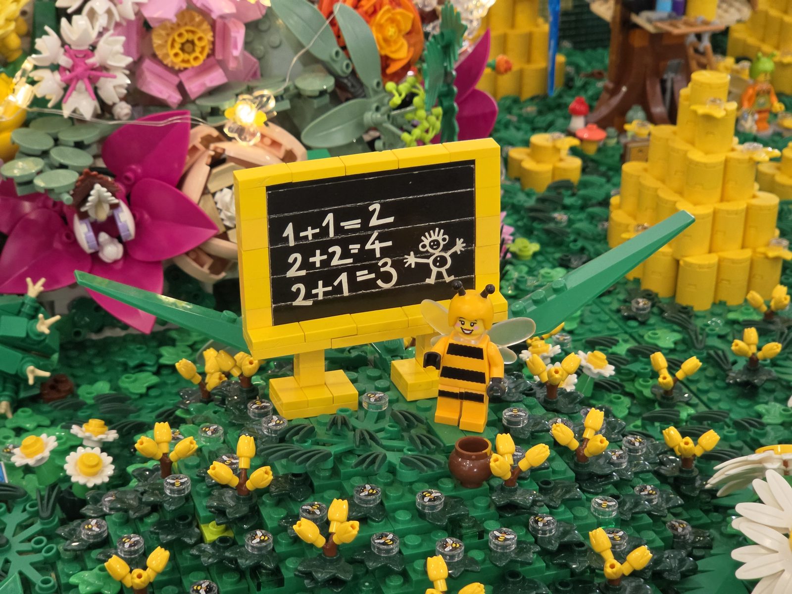 Eine kleine Tafel aus LEGO in einer Blumenwiese, darauf drei Rechenaufgaben und ein gemaltes Strichmännchen; davor eine Minifigur im Bienenkostüm